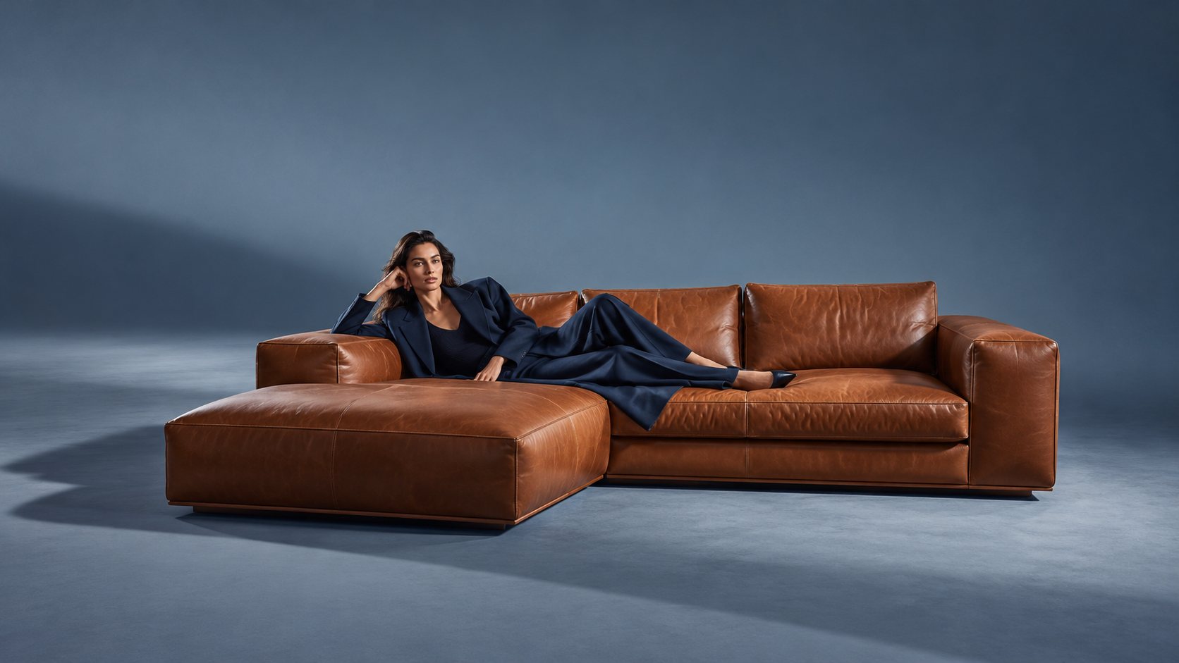 A model reclining on a cognac leather sectional against a deep blue backdrop